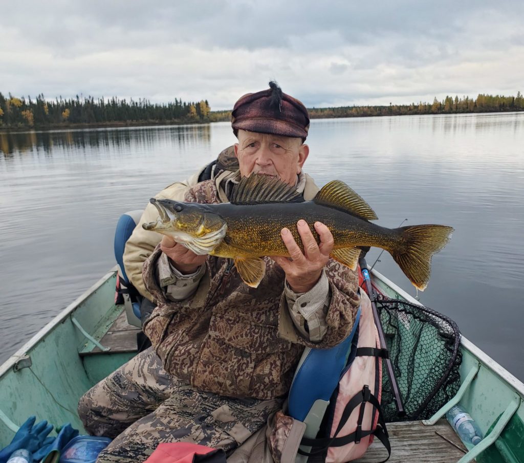 Gene holding a large walleye while sitting in a boat on water wearing their hunting gear